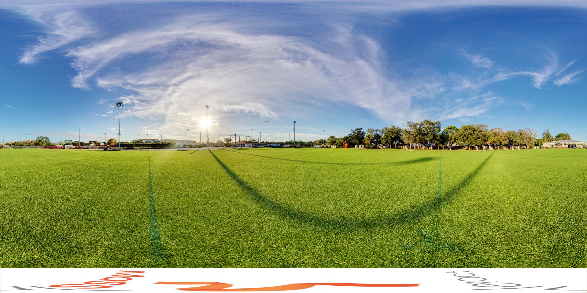 Panoramic view of a well-maintained soccer field under a vast blue sky with wispy clouds, featuring multiple floodlights and surrounded by trees and facilities